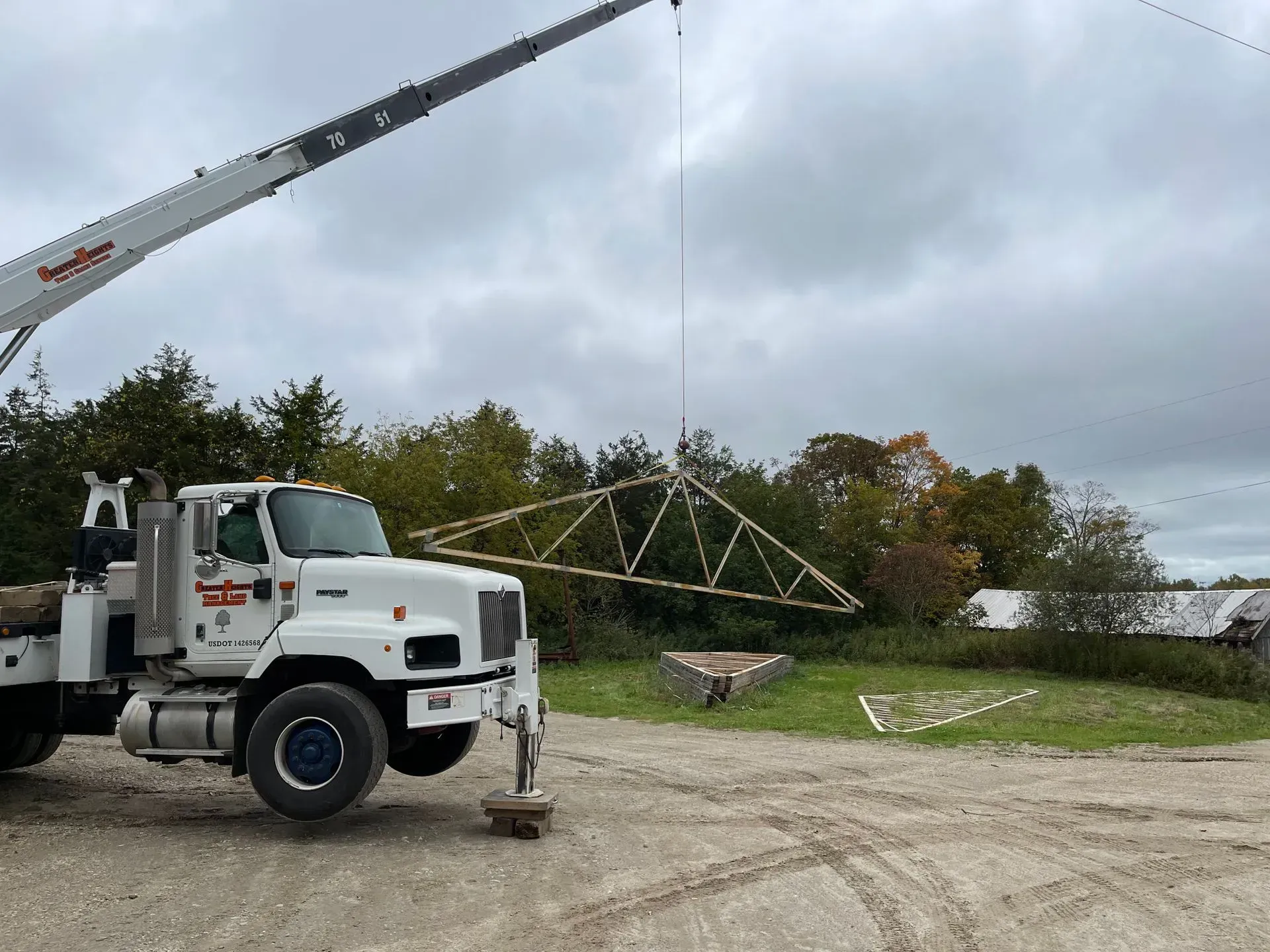 A white crane truck lifting a long, metal truss structure outdoors against a cloudy sky.