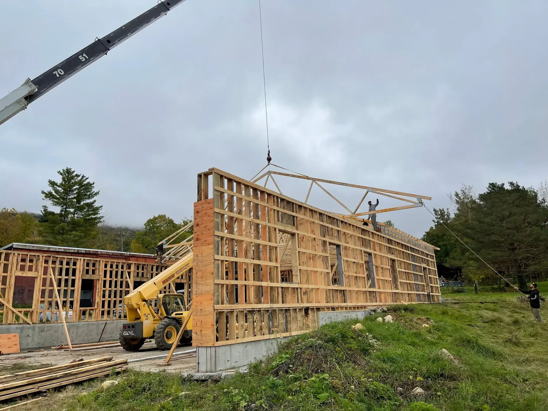 A construction crane lifts a roof truss onto the timber frame of a building under construction on a cloudy day.