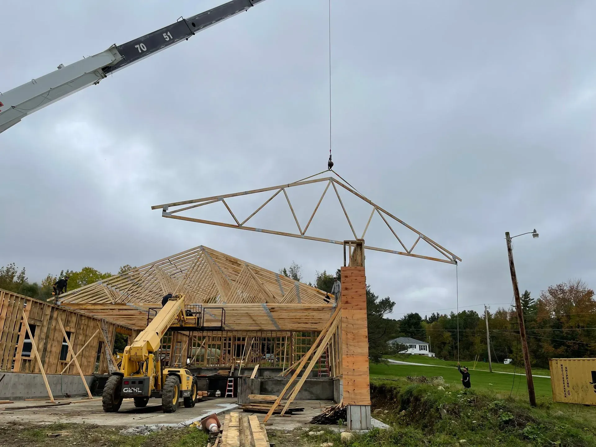 A crane lifts a wooden roof truss over a building under construction on an overcast day.