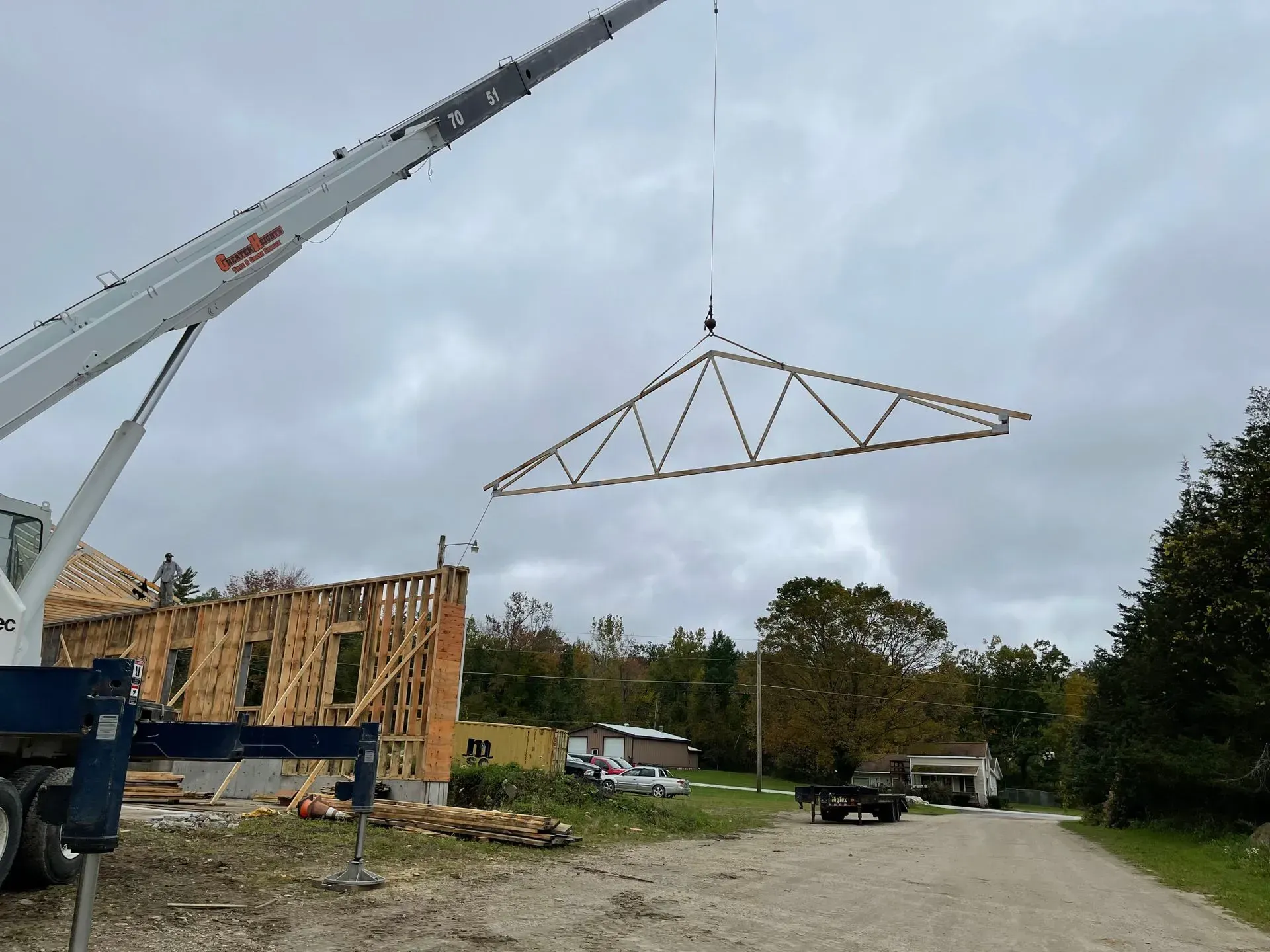 A crane lifting a wooden roof truss over a construction site on an overcast day.