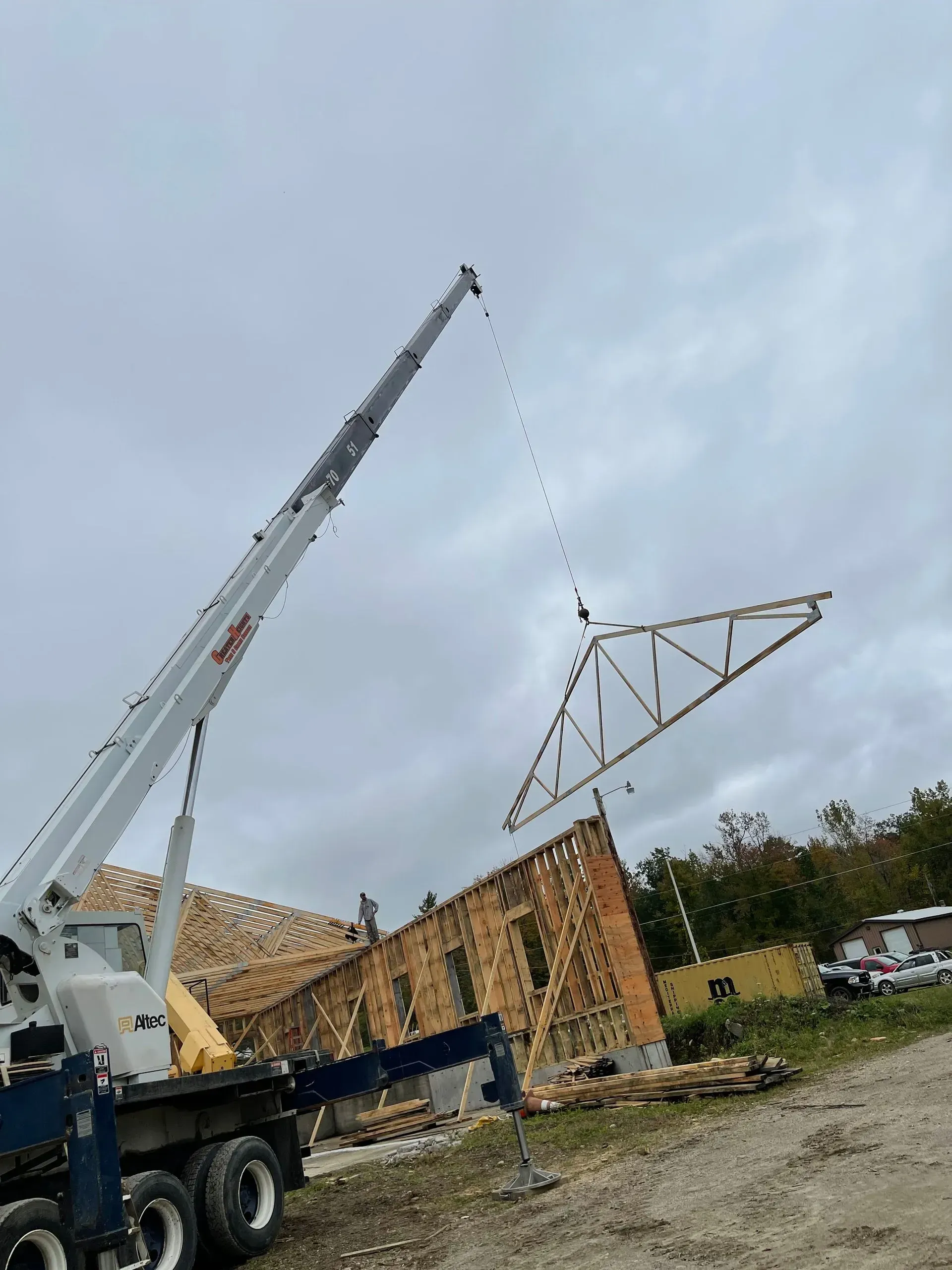 A crane hoists a wooden roof truss over the construction site of a building frame under a cloudy sky.