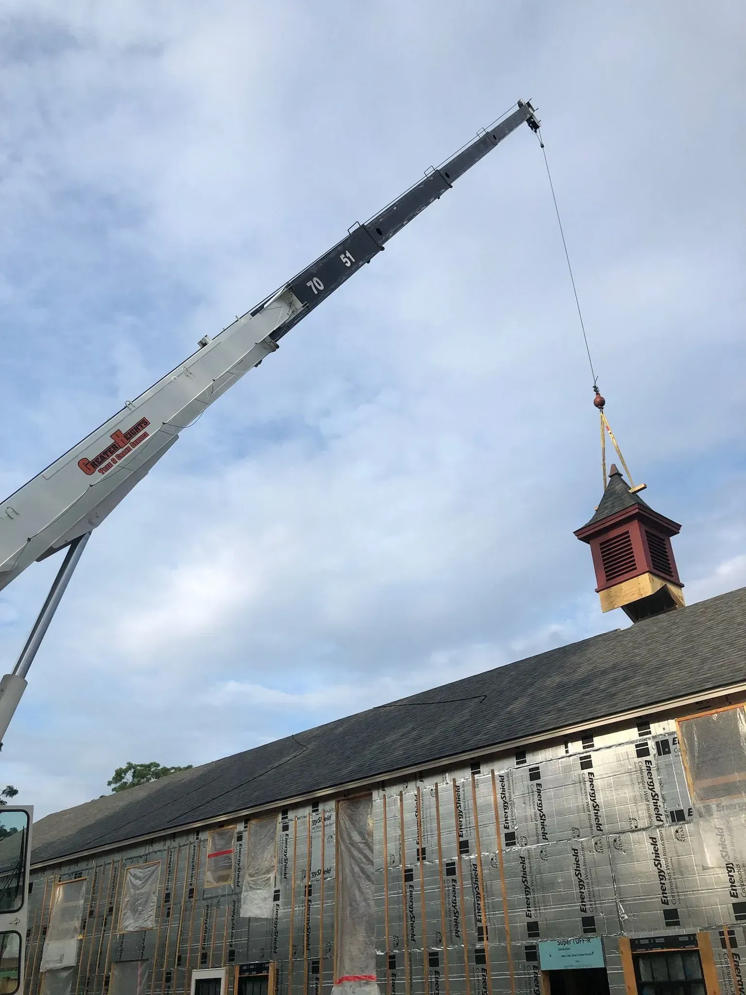 A large crane lifts a small, red-roofed wooden cupola into place atop a building under construction.