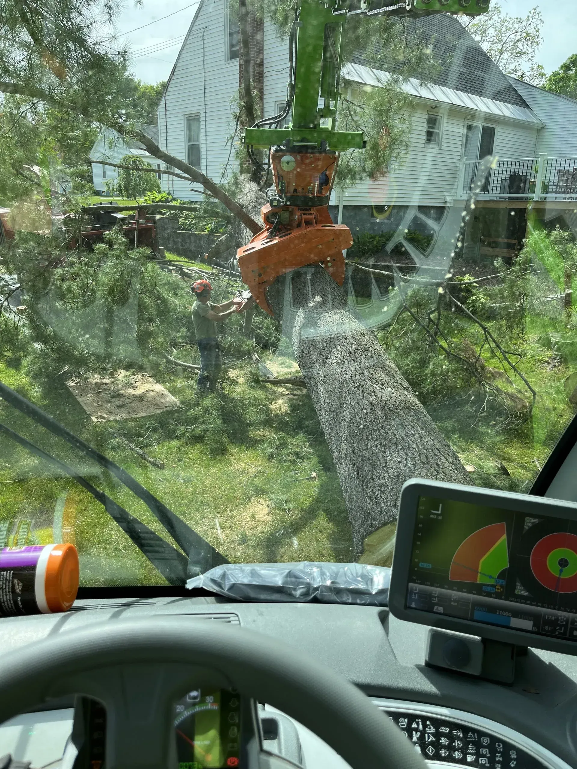A tree being cut with a mechanical arm, viewed from inside the cab of the operating vehicle, near a house.