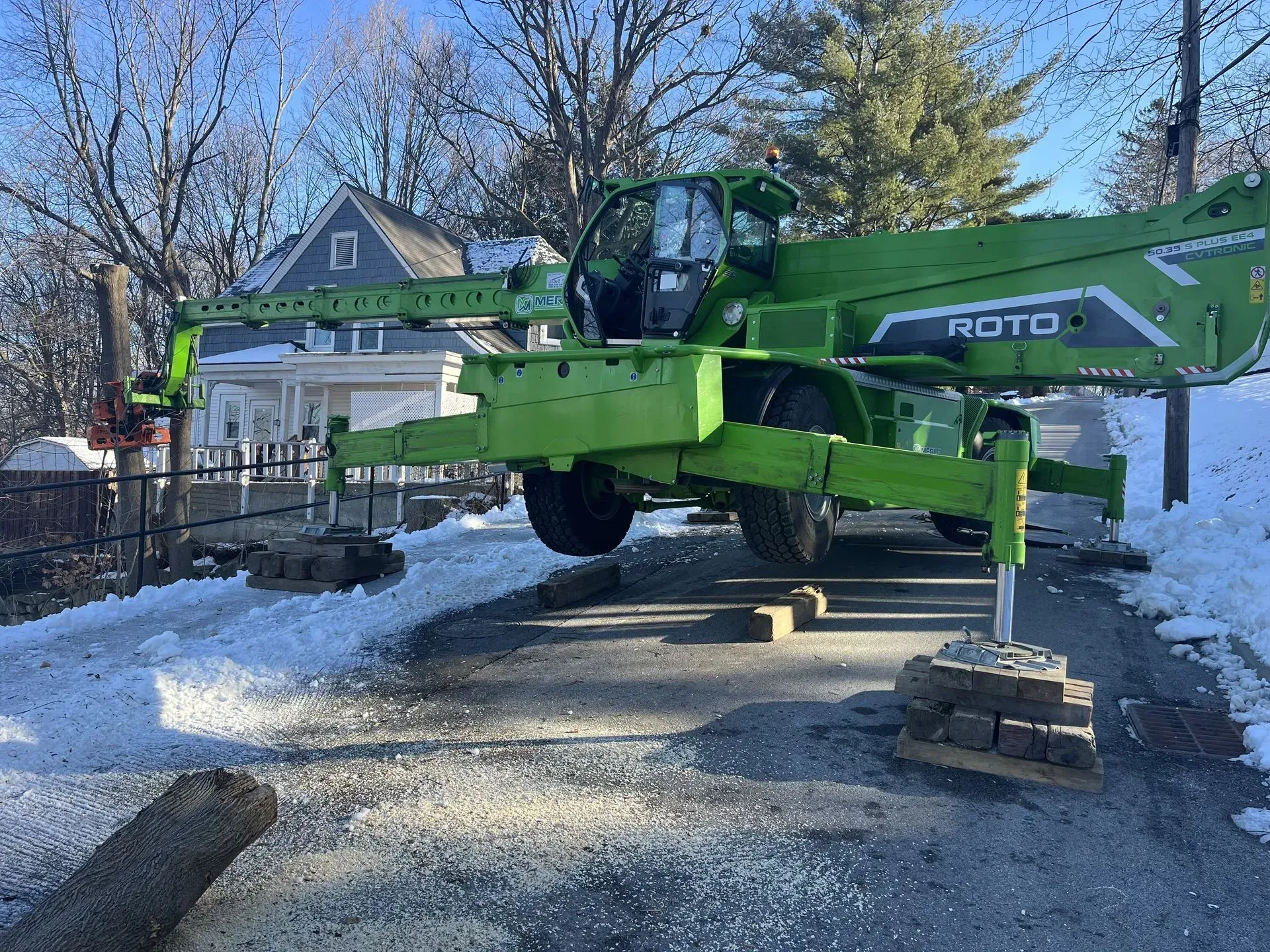 Green telehandler machine on a snowy driveway, supported by blocks. A house is in the background.