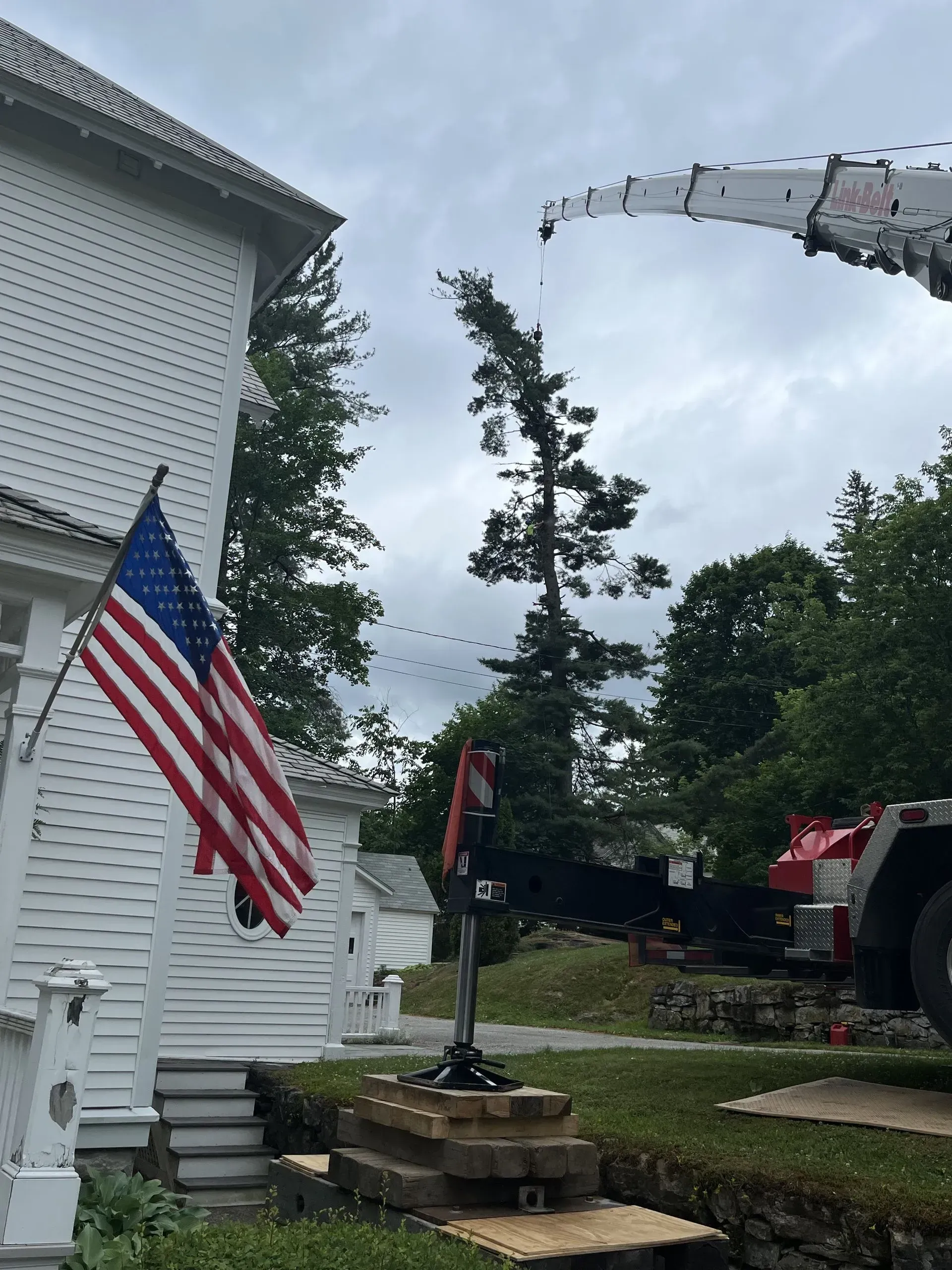 A crane lifts a tree section next to a white house with an American flag displayed on the porch.