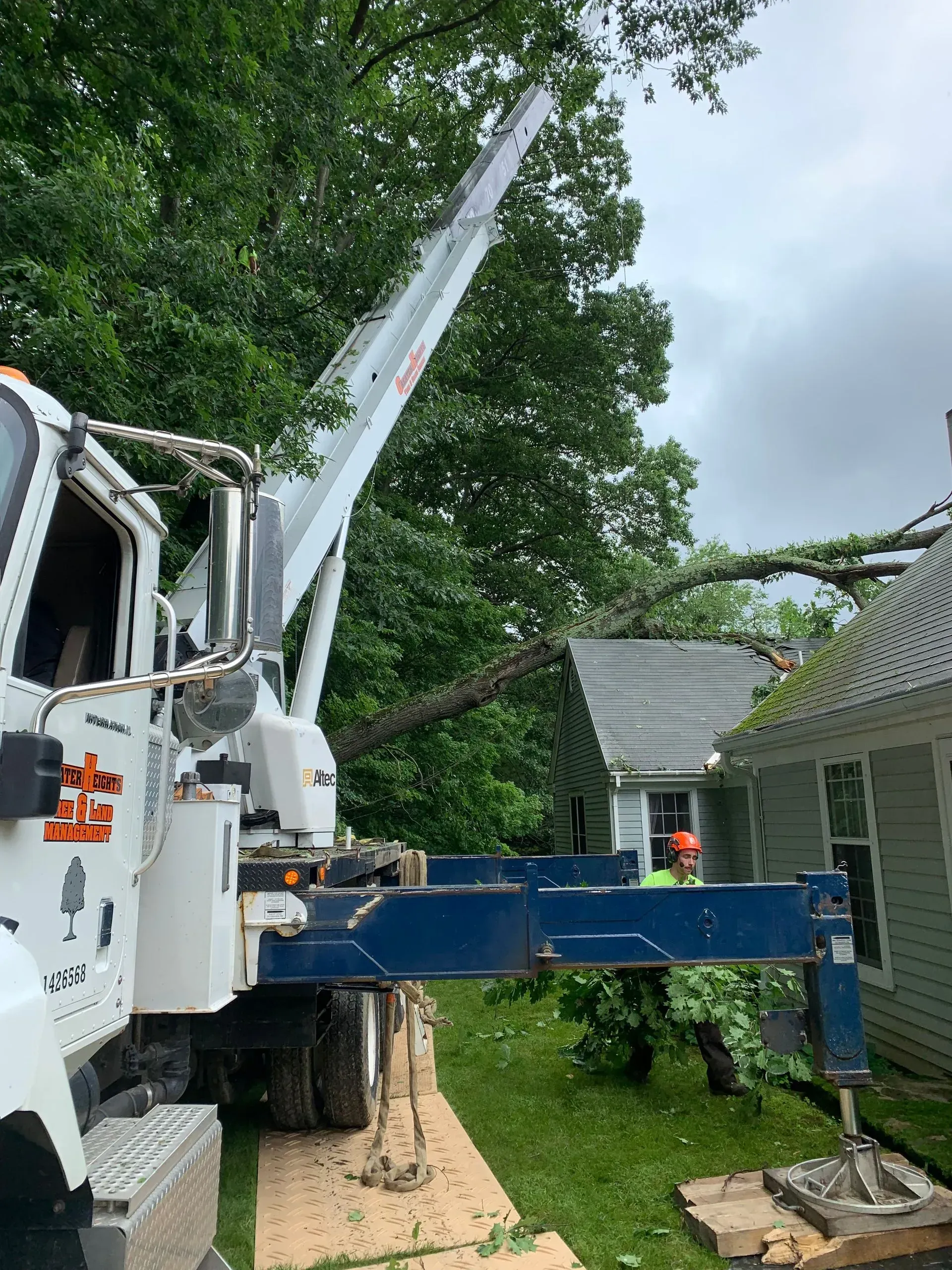 A white crane truck parked on a lawn, extending its boom over a house to remove a fallen tree.