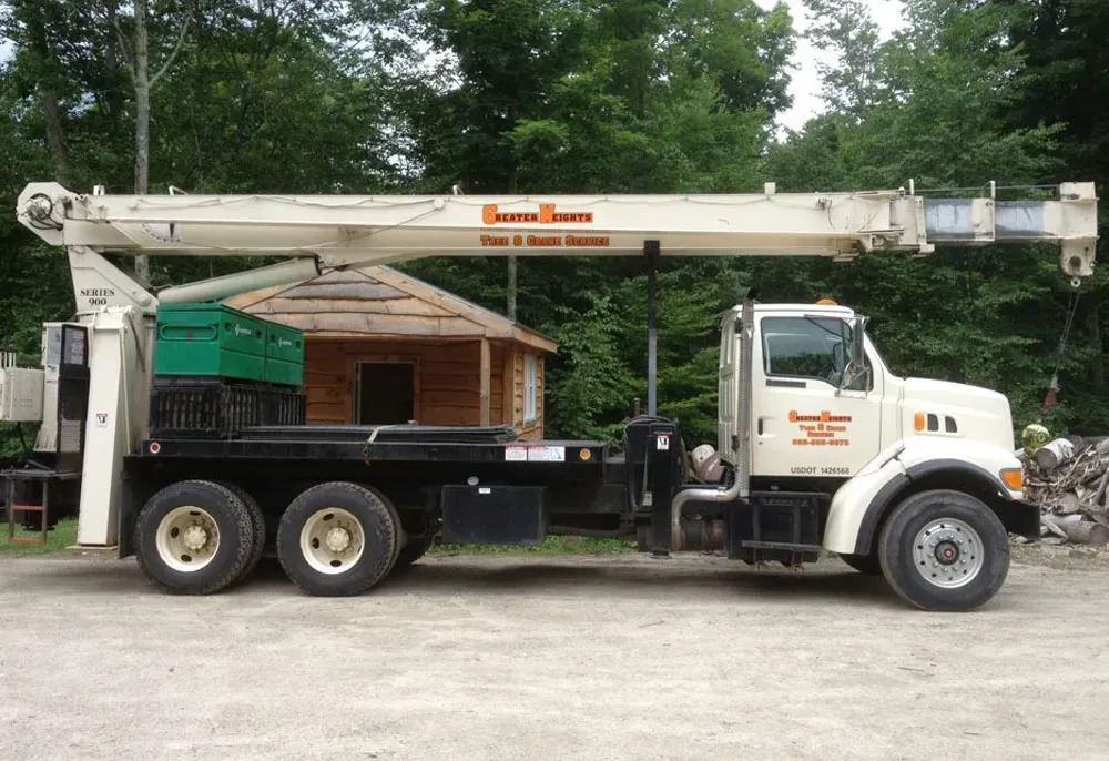 A large white boom truck with a crane is parked outdoors near a small wooden building and trees.