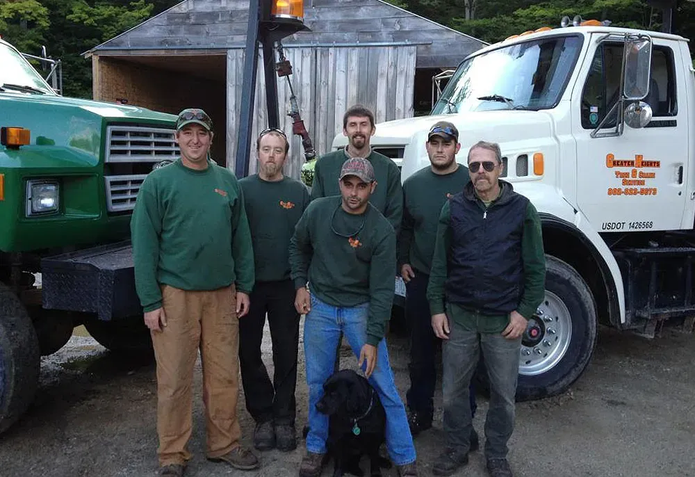 Six men and a black dog posing with two work trucks in front of a shed.