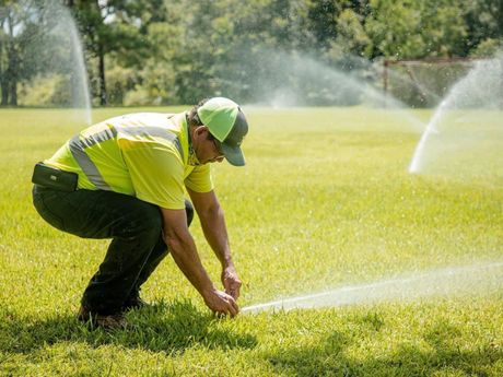 A man is kneeling down on a lush green field to adjust a sprinkler.