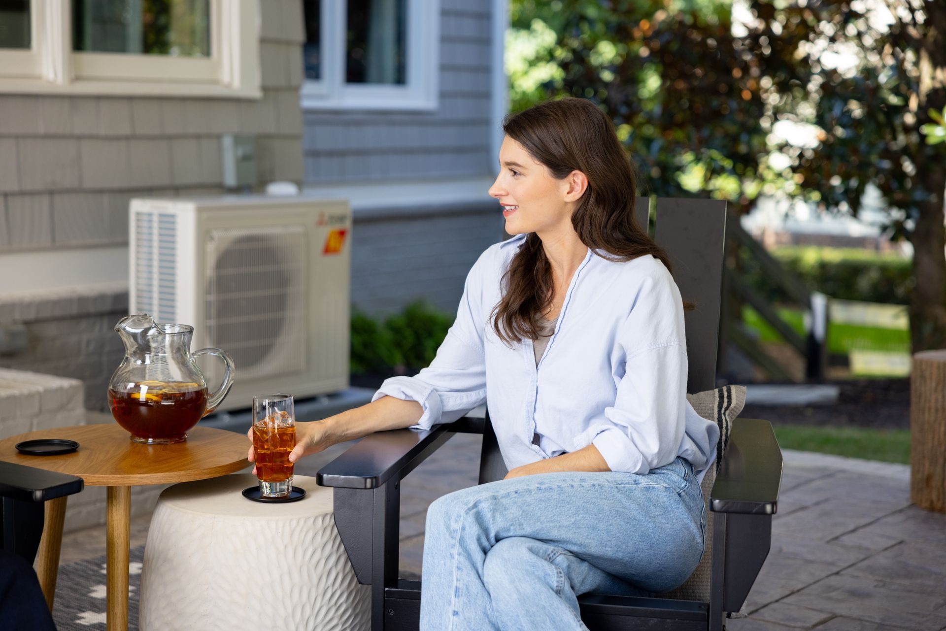 Woman sitting outside, holding a drink. She's smiling, near a table with a pitcher of iced tea and air conditioning unit.