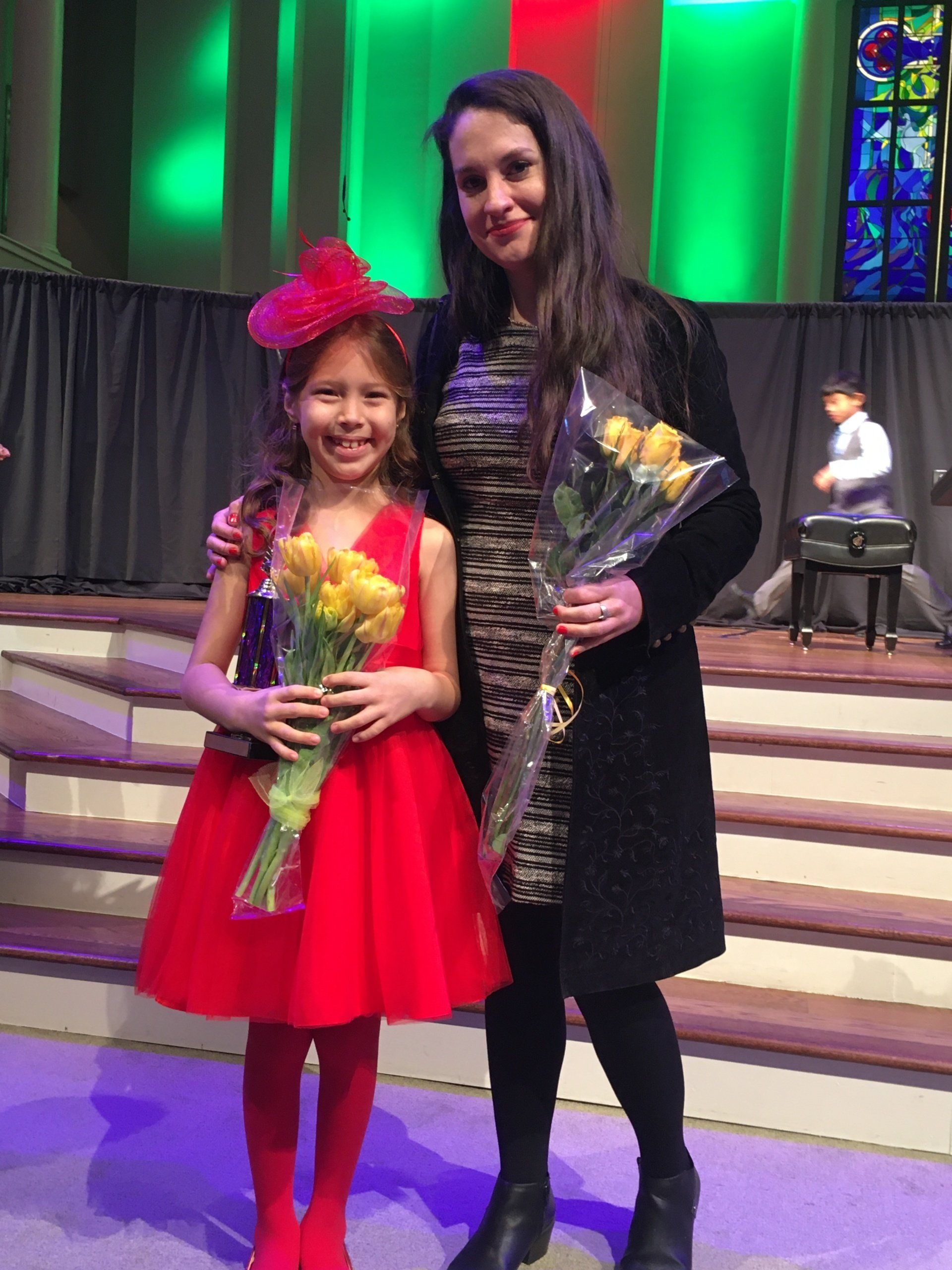 Girl in red dress and hat with woman holding flowers on stage.