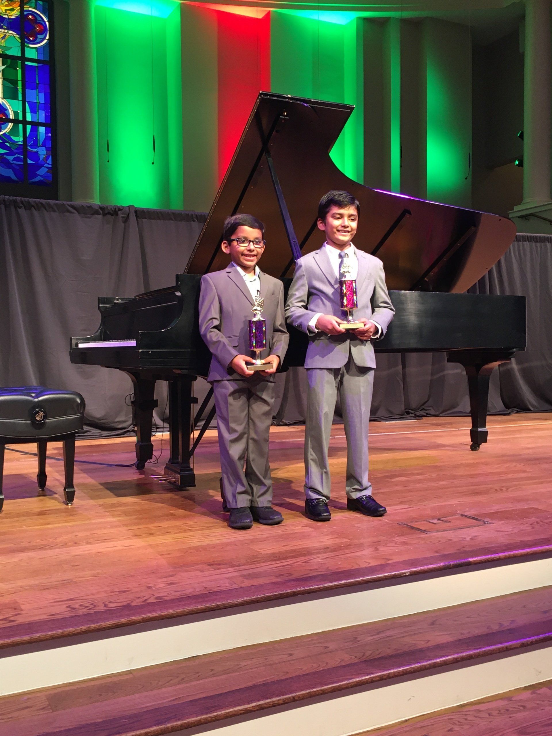 Two boys in suits holding trophies stand on a stage in front of a grand piano.