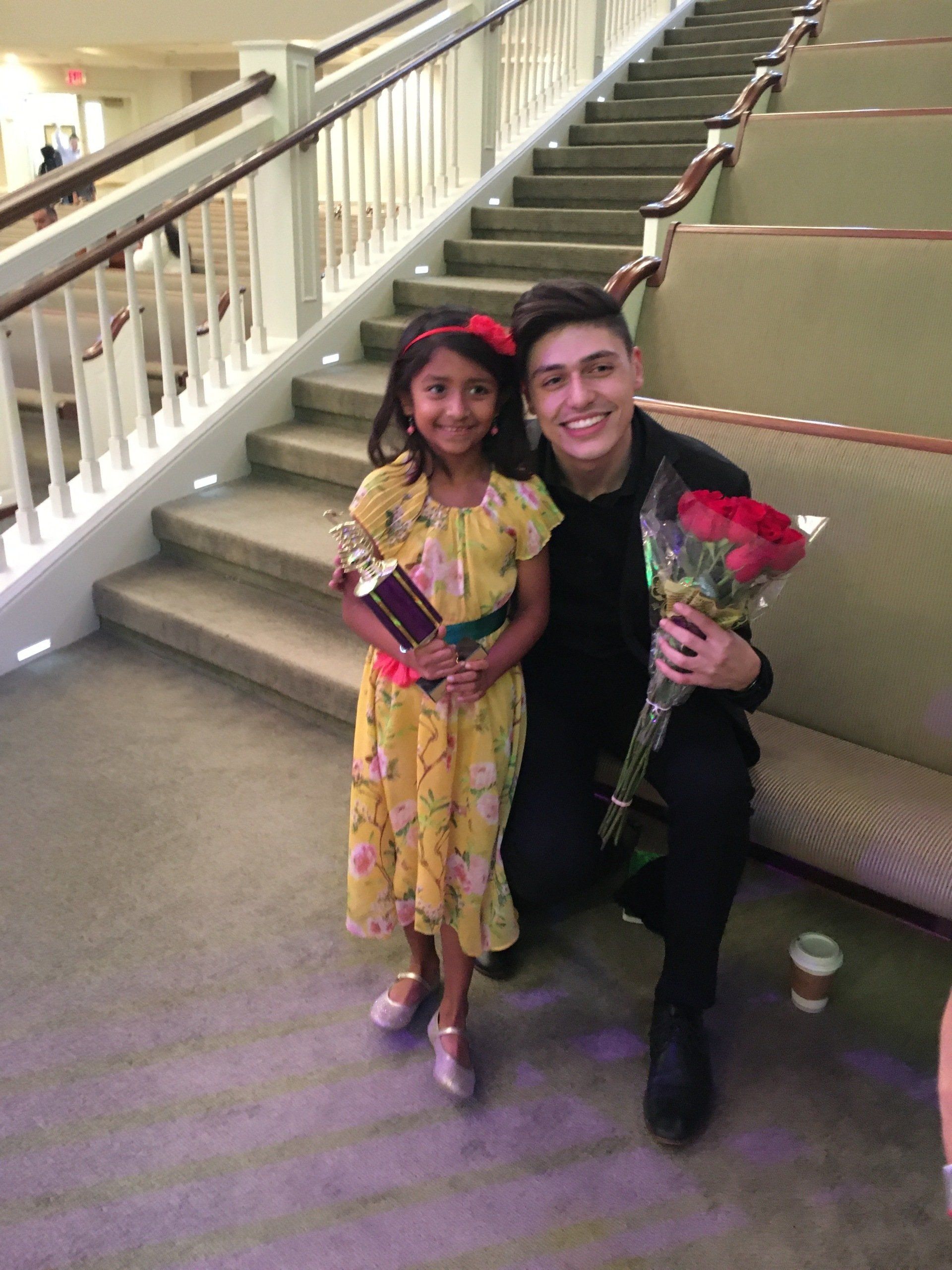 A young girl in yellow dress and a man holding roses smile on a staircase.