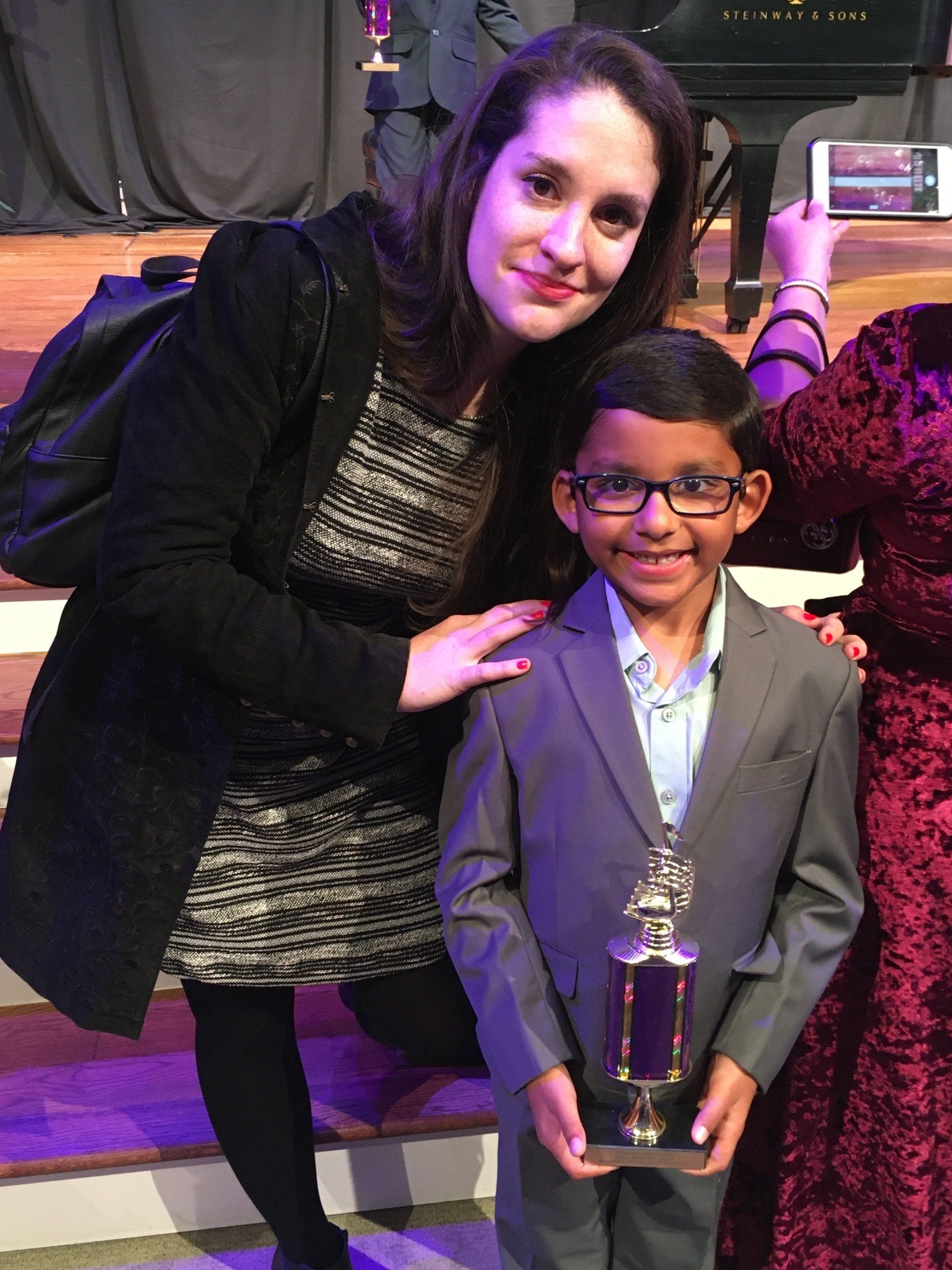 Woman and boy in a suit smiling and holding a trophy on a stage, possibly at an awards ceremony.