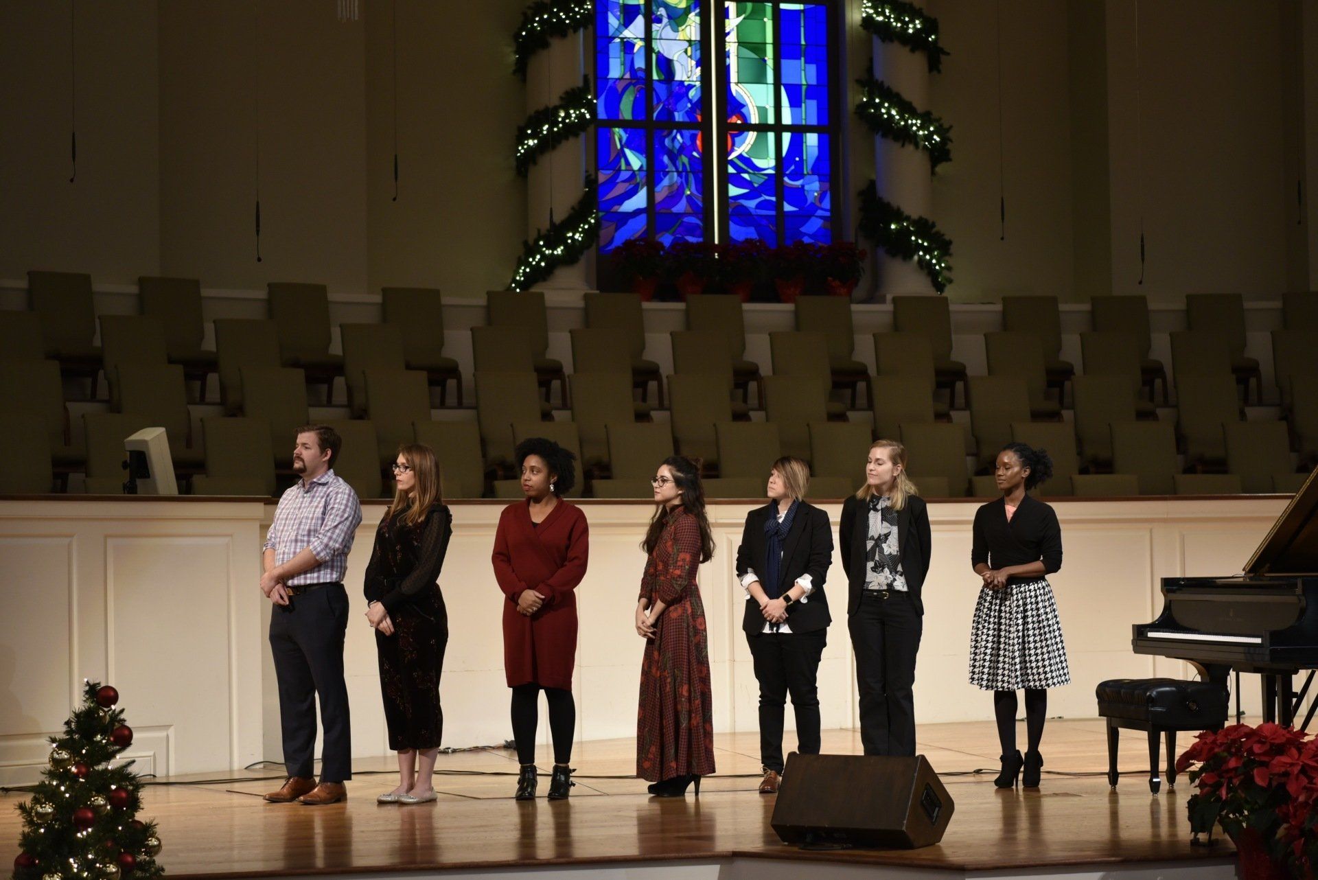 Choir on stage in a church, standing in front of a stained-glass window, with a piano on the right.