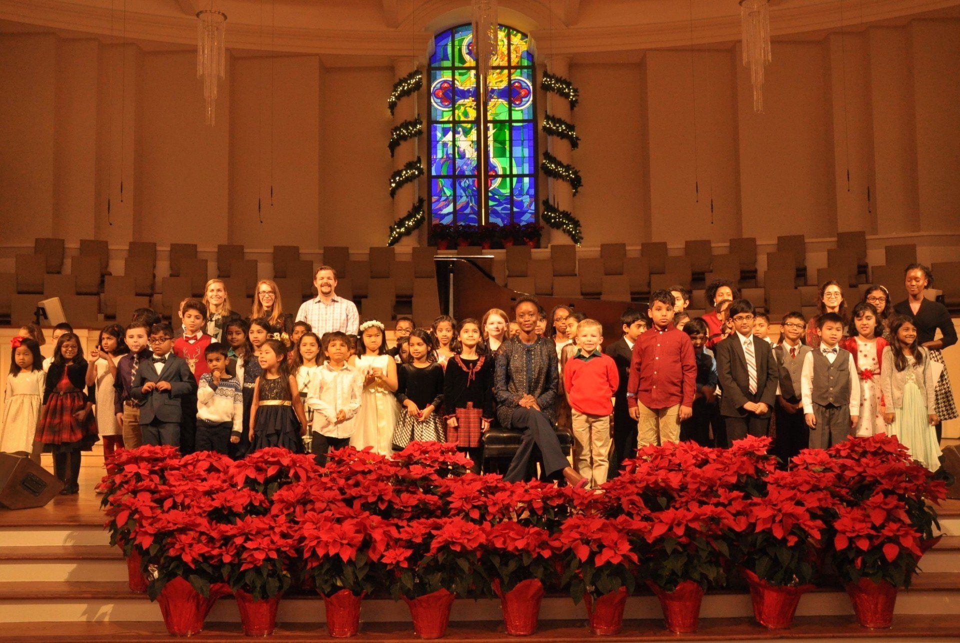 Children's choir with director on stage, surrounded by poinsettias in front of a stained-glass window.