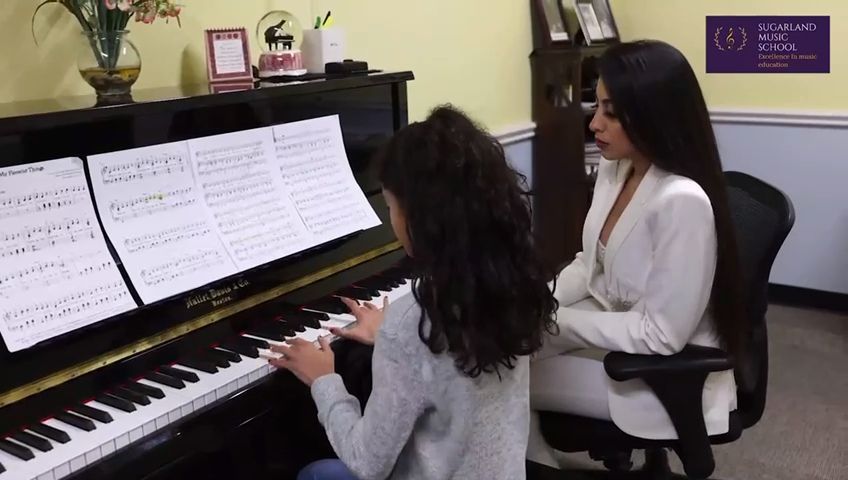 Young girl playing piano with a woman seated beside her in a music room.