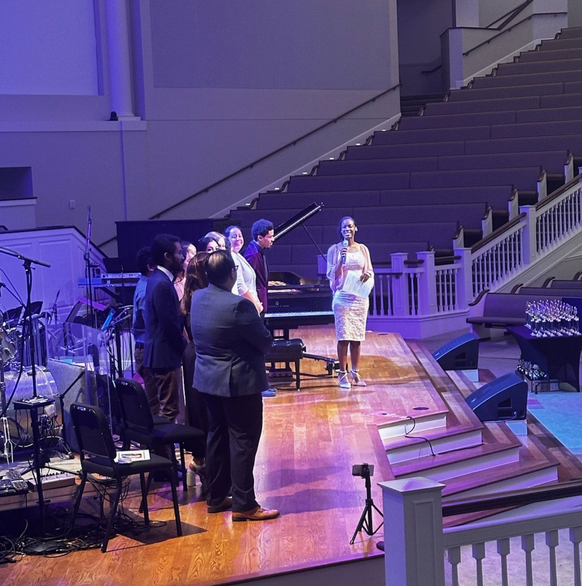 People on stage, a woman speaking, a piano, audience seating, purple lighting.