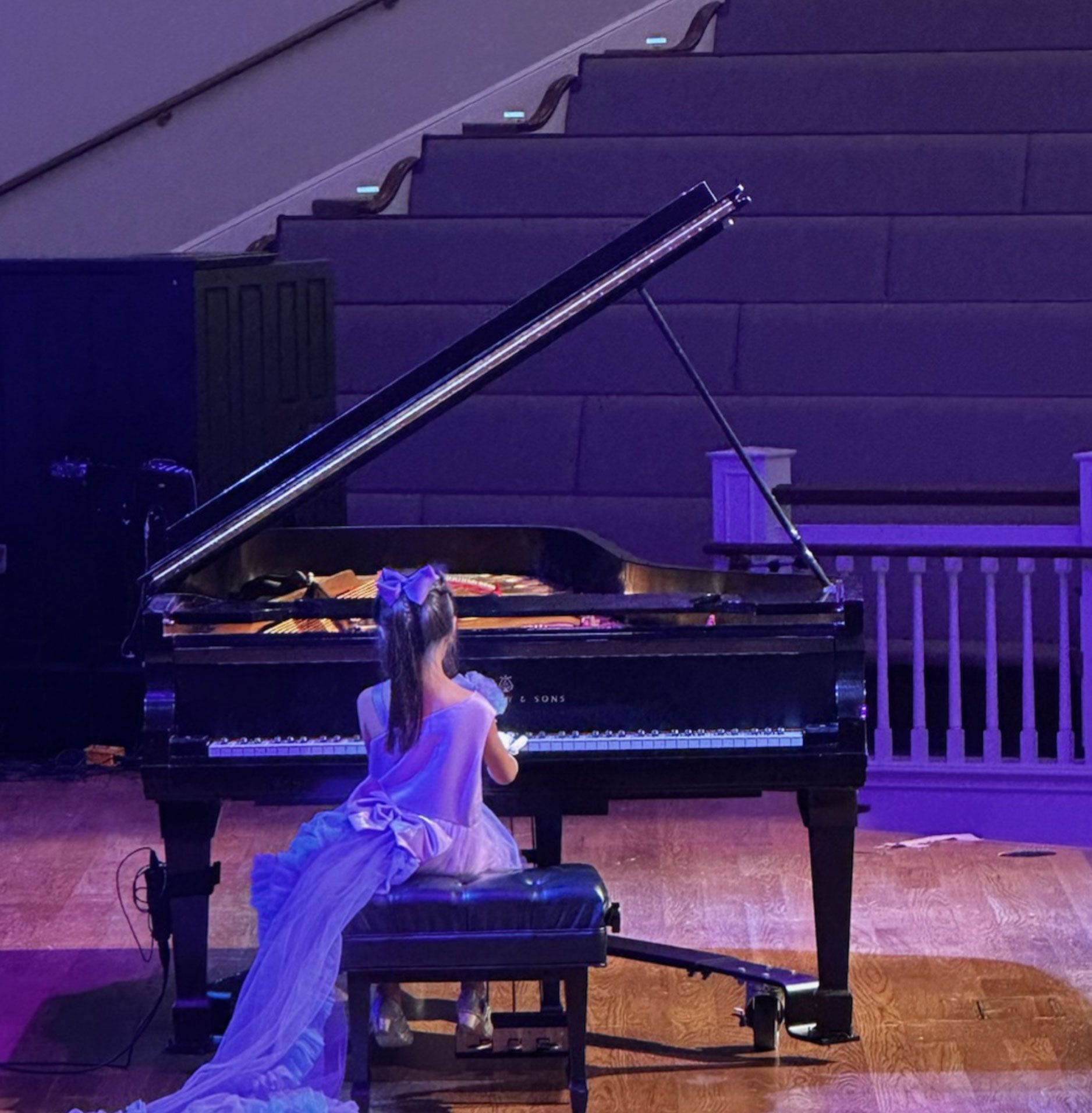 Young girl in a purple dress plays a grand piano onstage.