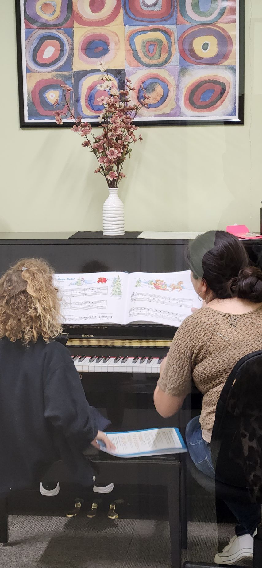 Two people at a piano, music sheets on the stand, one playing. A colorful artwork is on the wall.