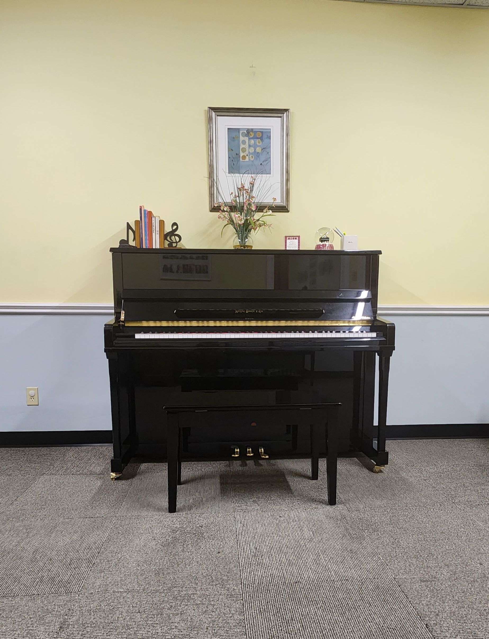 Black upright piano with bench on patterned carpet, against a pale yellow wall with artwork.