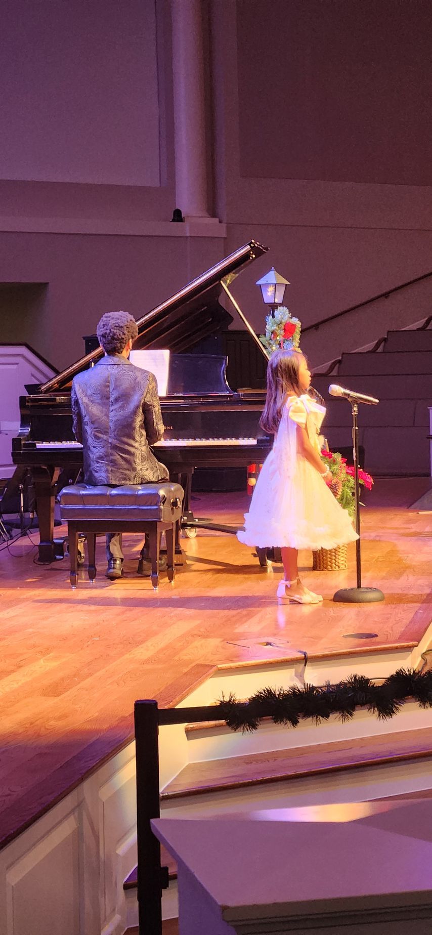 A young girl in a white dress sings into a microphone on stage with a pianist at a grand piano.
