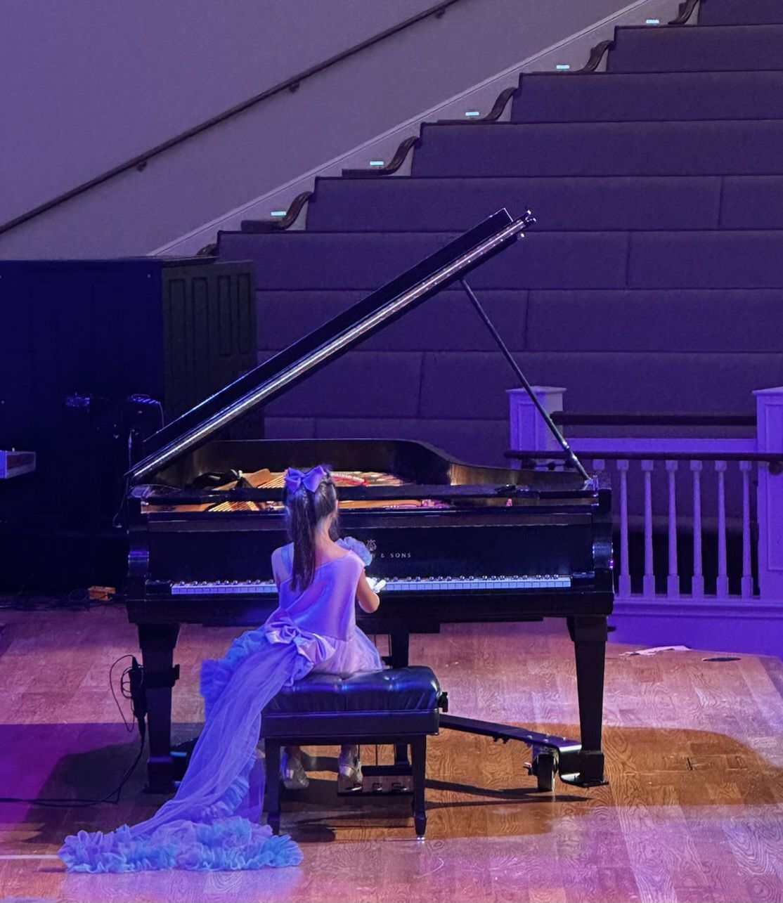 Young girl in a purple dress playing a grand piano on a stage with stairs in the background.