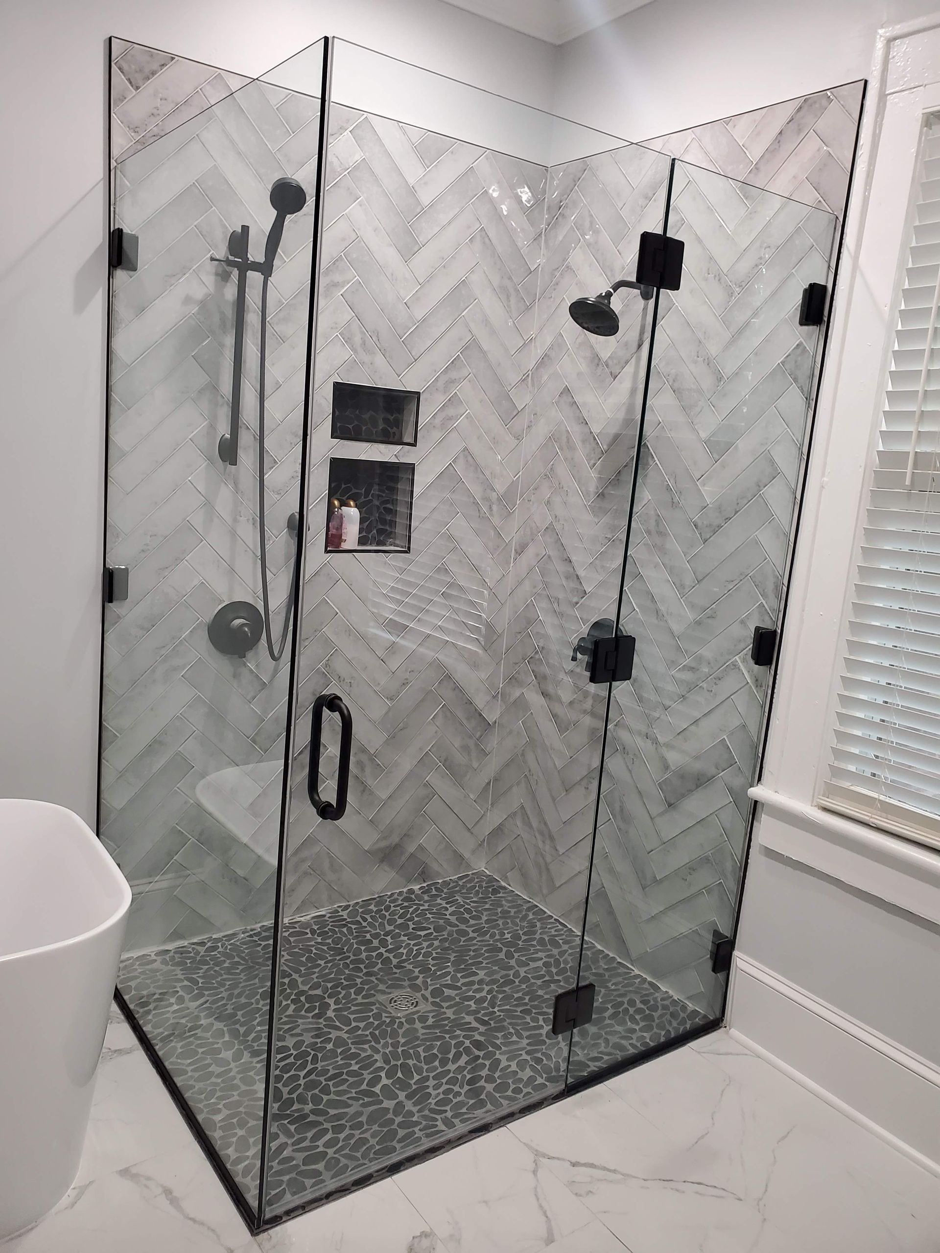 Corner glass shower with gray herringbone tile and black fixtures in a bathroom