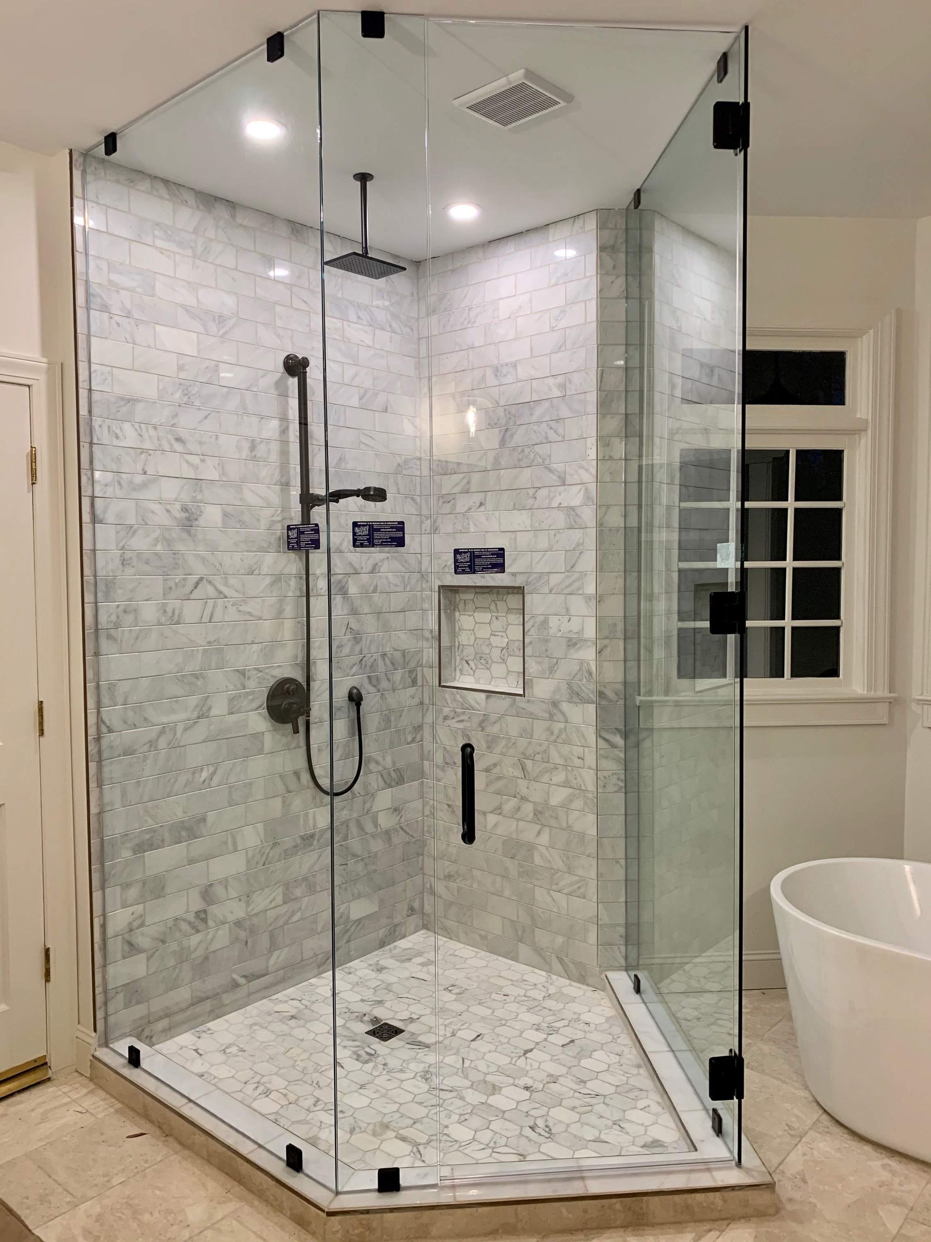 Modern glass-enclosed corner shower with marble tile and brushed metal fixtures in a bright bathroom