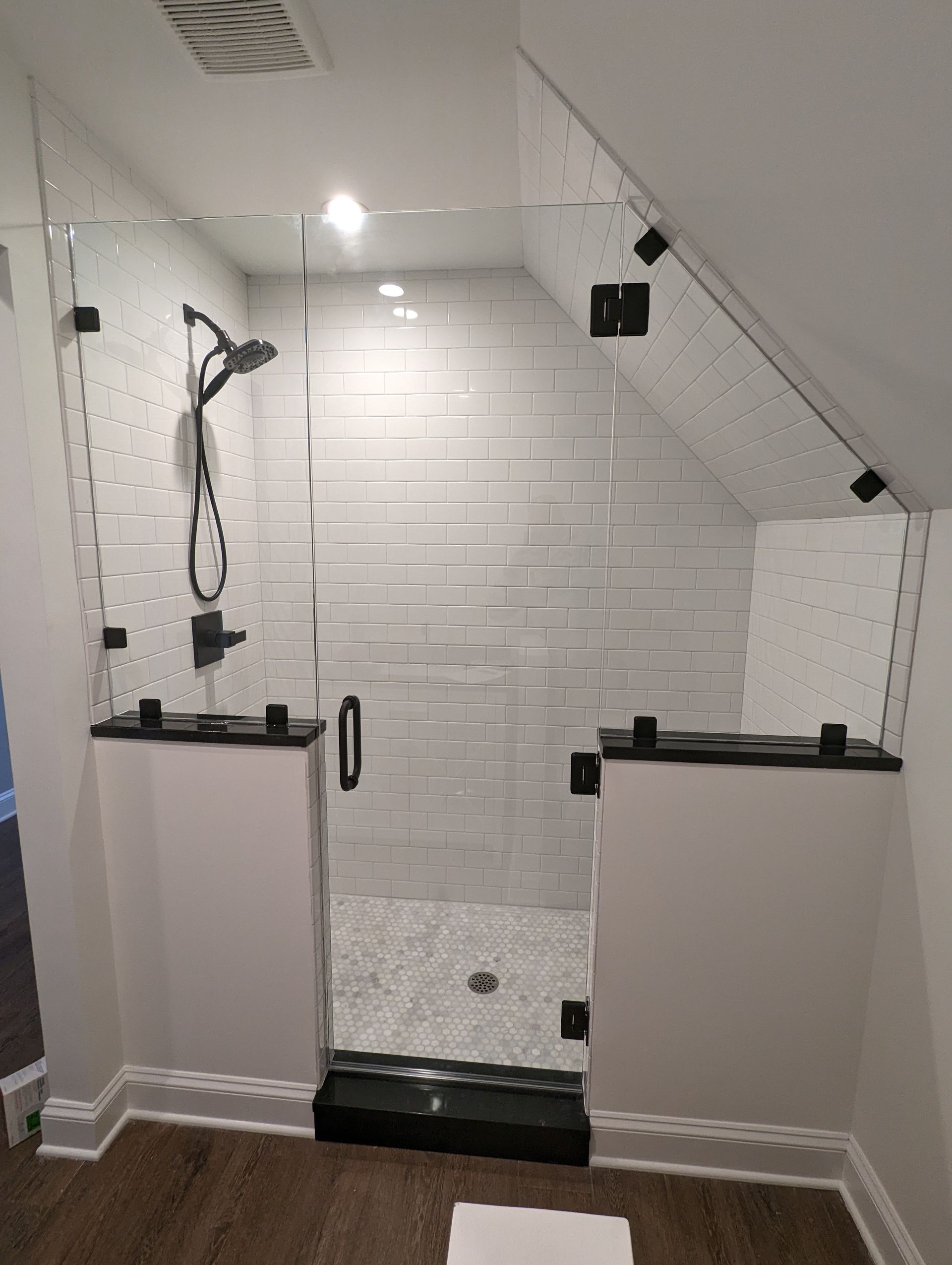 White tiled shower with handheld showerhead and glass doorway under a sloped ceiling