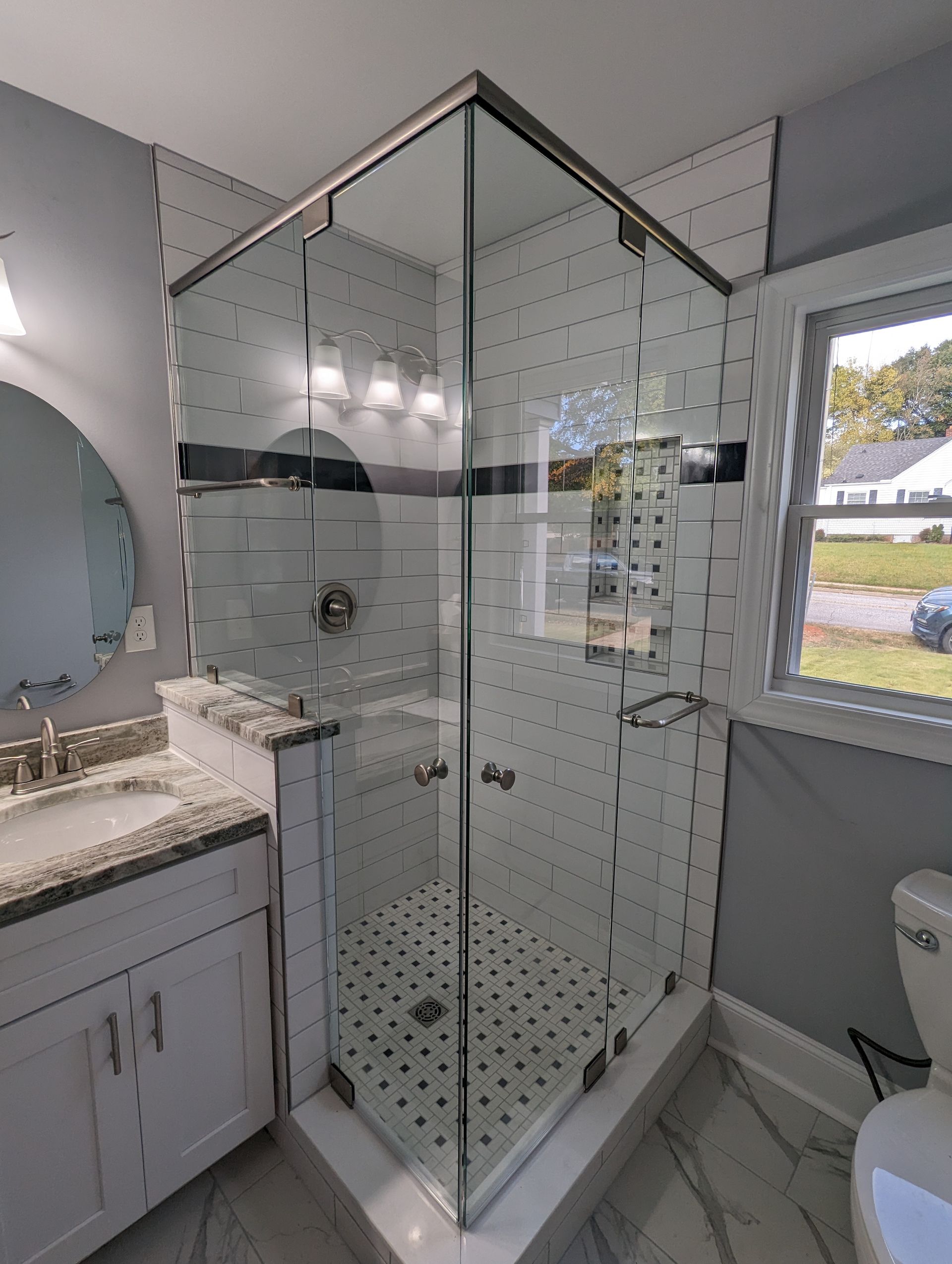 Modern bathroom with glass shower, white vanity, round mirror, and window view of greenery.