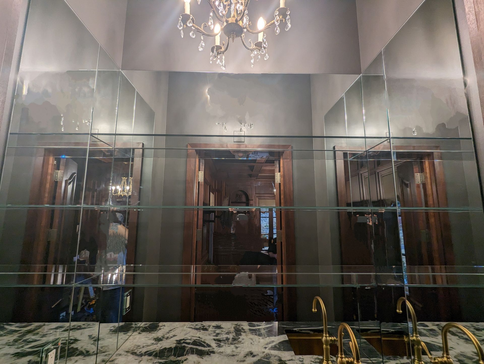Elegant mirrored bathroom with a chandelier, dark cabinetry, and a gold faucet over a black marble countertop