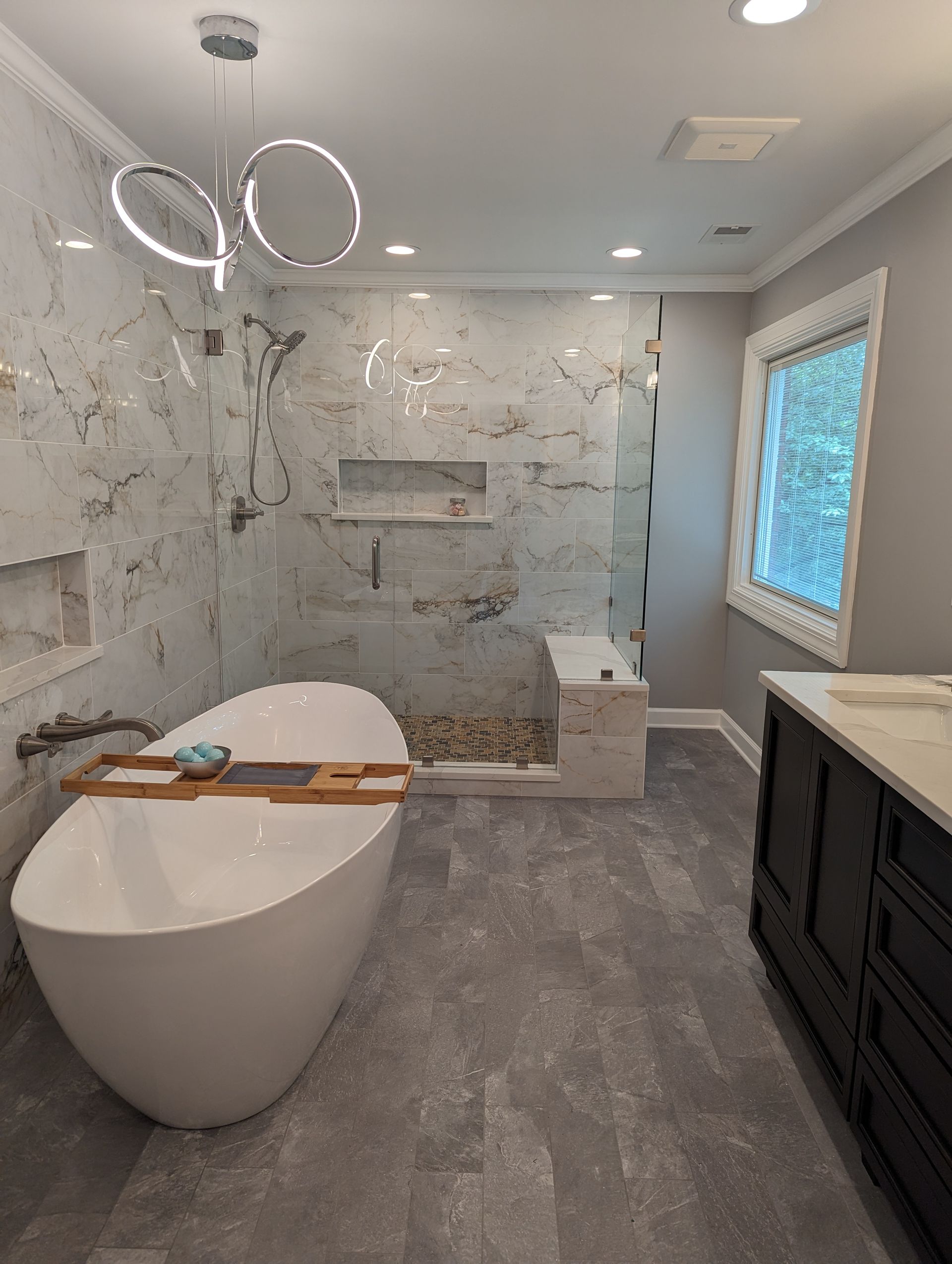 Modern bathroom with freestanding tub, glass shower, gray tile, and dark vanity near a window