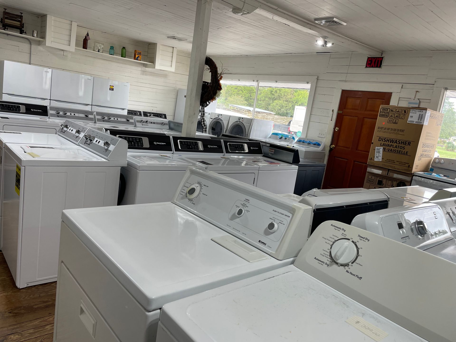 Rows of white and black washing machines and refrigerators in a store, with a brown door and windows visible.