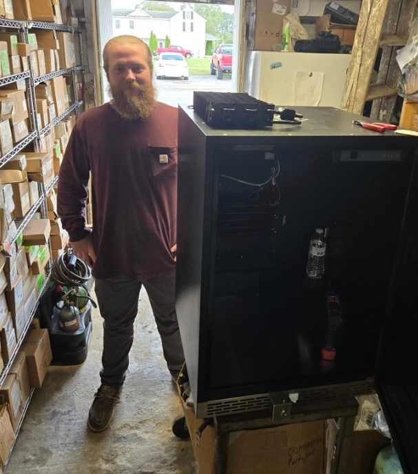Man with beard stands next to black server cabinet in cluttered storage area.