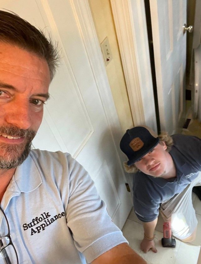 Man taking a selfie with a younger man installing appliance. Both are in a doorway. The man has a company logo on his shirt.