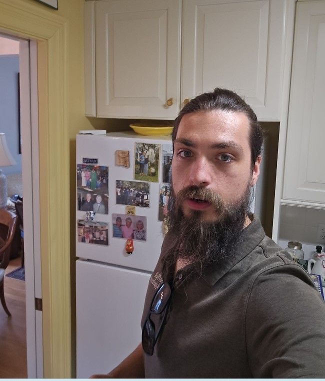 Man with beard in a kitchen, taking a selfie. Wearing a green shirt, standing in front of a refrigerator.