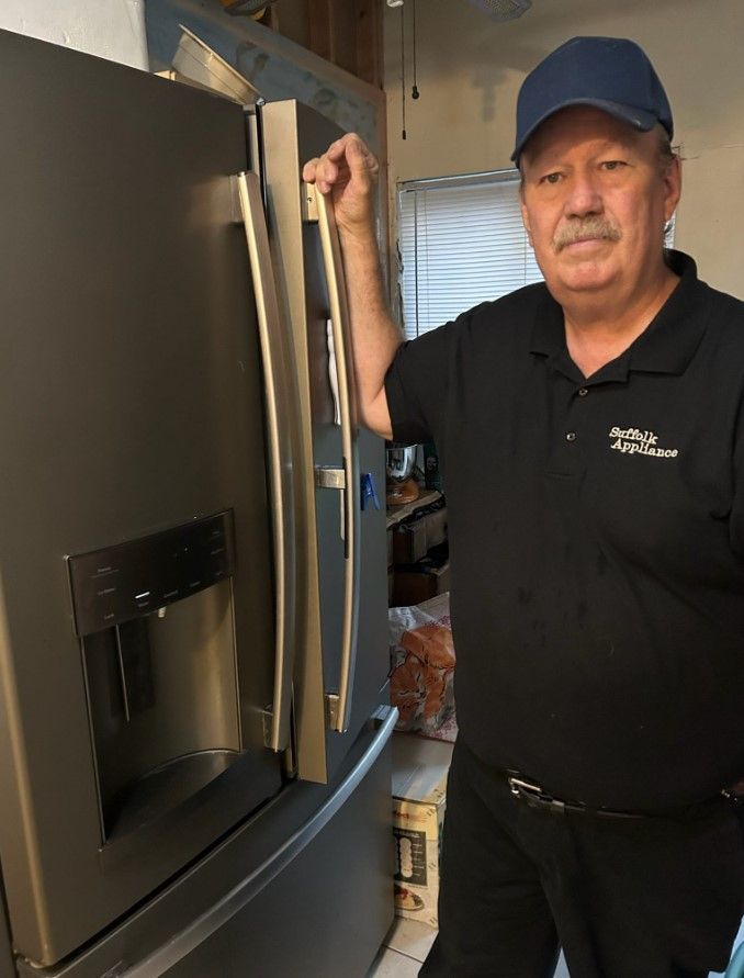 Man in black shirt and cap stands next to a dark gray refrigerator, hand on the door.