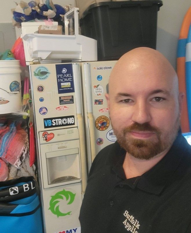 Bald man with beard, standing in front of a refrigerator with stickers on it, Suffolk Appliance shirt.