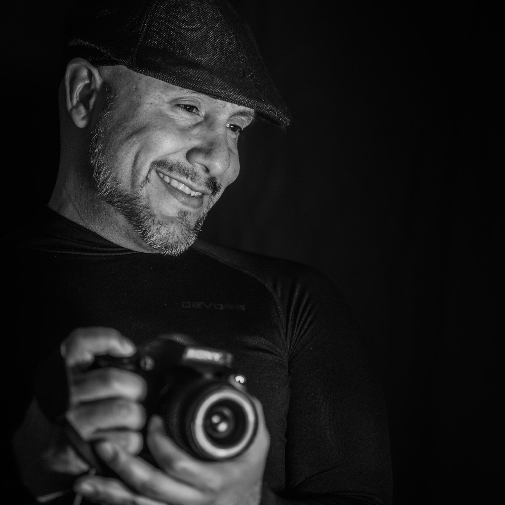 Man smiling, holding camera, wearing cap, black background.