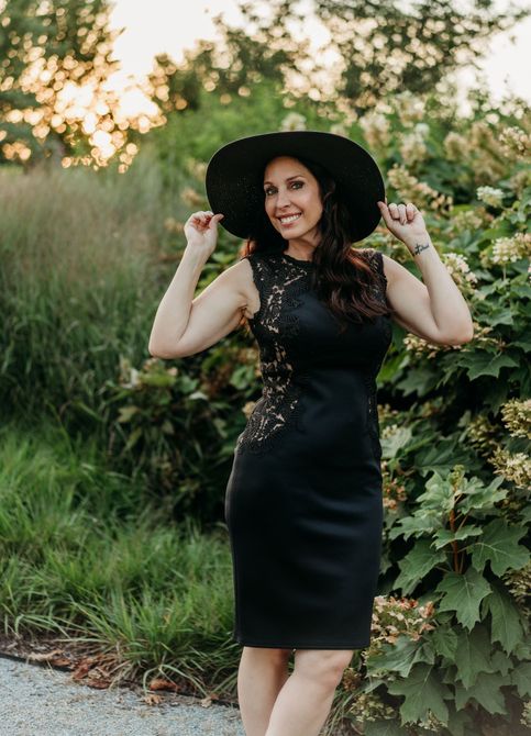 A woman in a black dress and hat is standing in front of a bush.