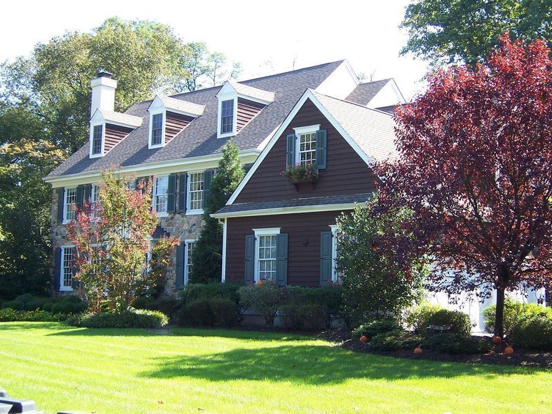 Two-story brown and stone house with dormers, green shutters, and a red tree in the front yard.