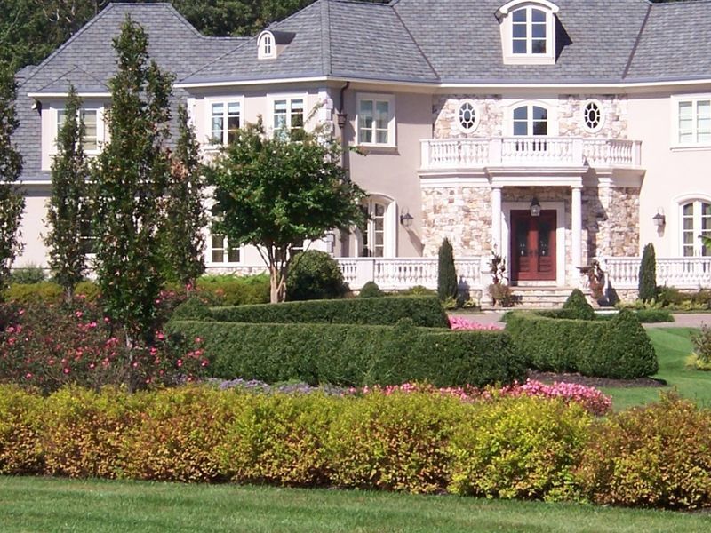 Large, light-colored house with a formal garden featuring manicured hedges, colorful bushes, and trees.