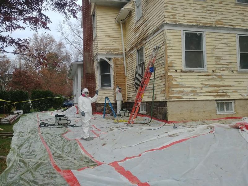 Workers in protective suits removing paint from a house; debris tarp, ladder.