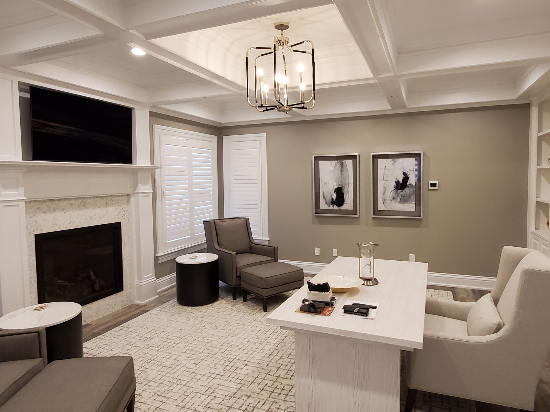 A bright home office with a fireplace, a white desk, two armchairs, and abstract wall art under a coffered ceiling.