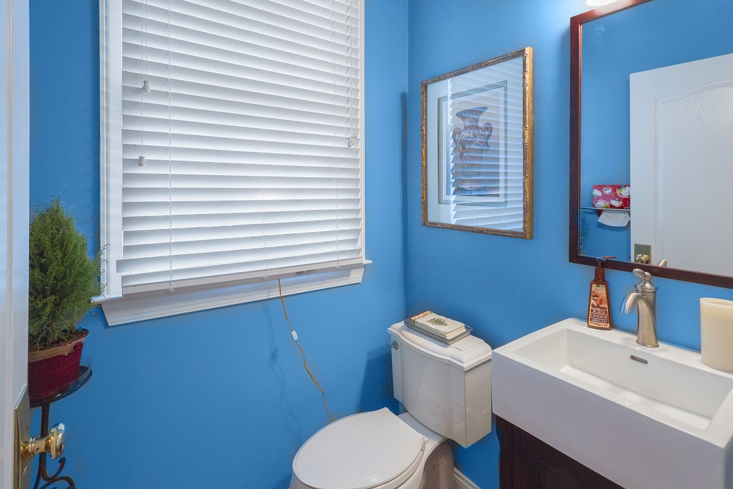 Blue bathroom with toilet, sink, mirror, and window blinds.