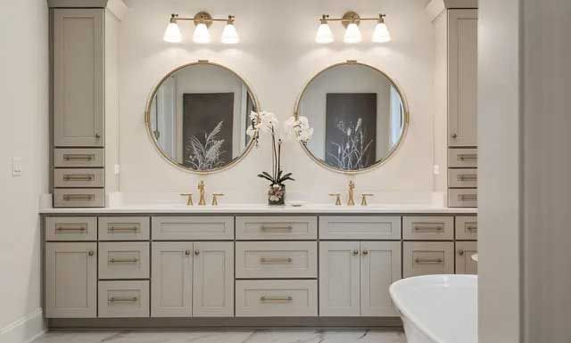 Bathroom with double vanity; two round mirrors, gold fixtures, gray cabinets, white countertops.