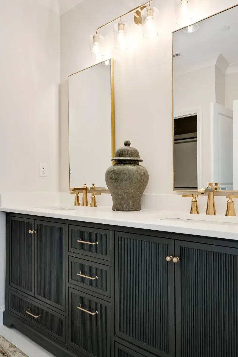 Dark green bathroom vanity with gold accents, white countertop, mirrors, and decorative jar.