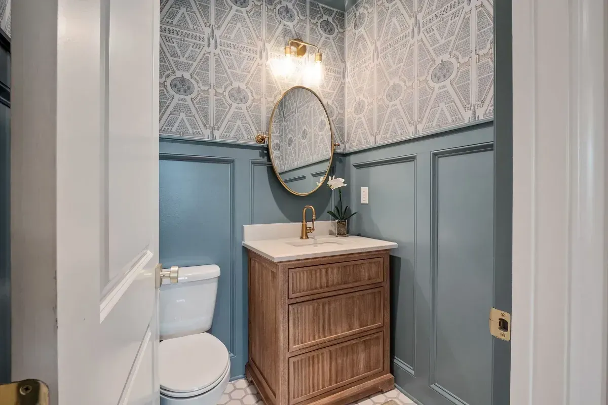 Powder room with blue wainscoting and patterned wallpaper; wood vanity, oval mirror.