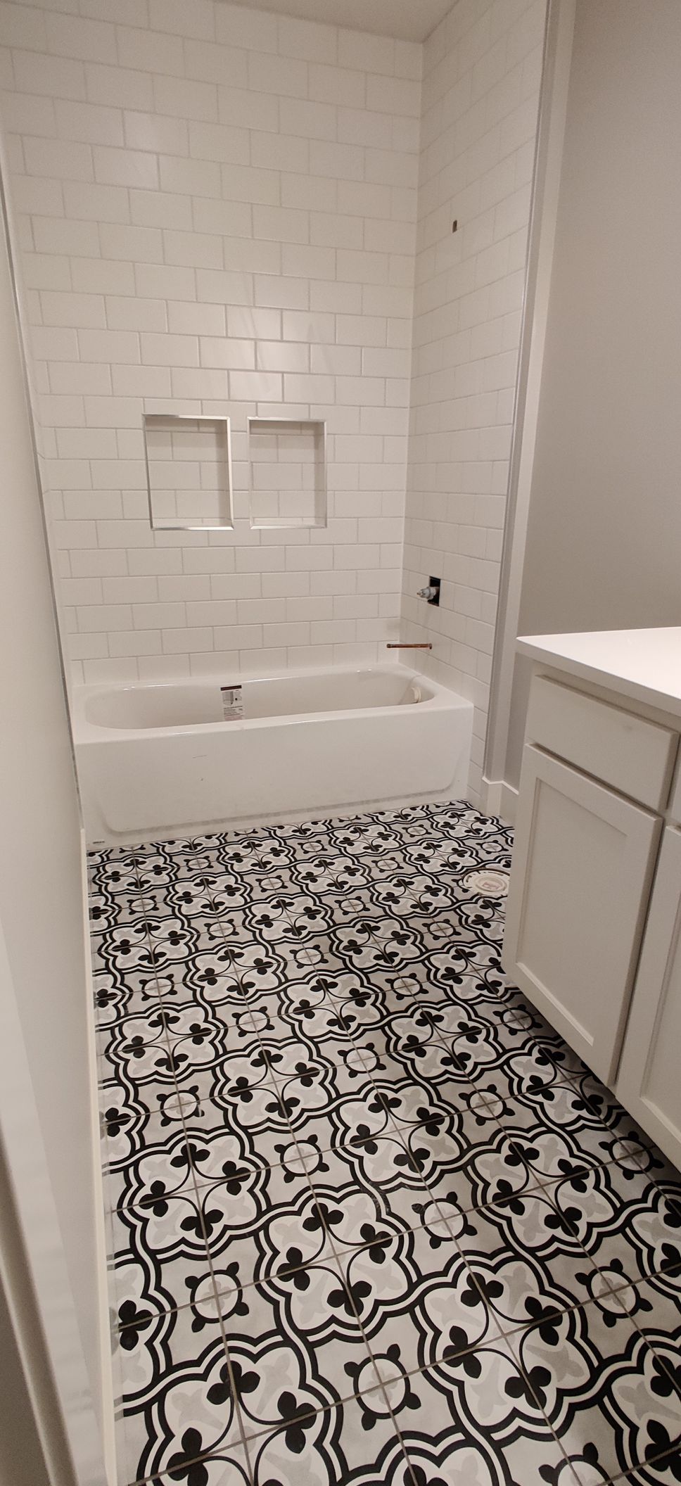 White bathroom with patterned black-and-white tile, tub-shower niche, and vanity cabinet.